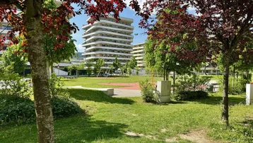 Grüne Außenanlage mit Spielplatz und modernen Wohngebäuden im Hintergrund unter blauem Himmel.