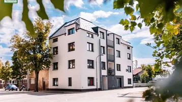 Modernes Mehrfamilienhaus mit weißer Fassade und dunklen Fenstern, umgeben von Bäumen und Straße.