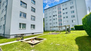 Zwei Mehrfamilienhäuser mit großem Gemeinschaftsgarten, Spielplatz und Sandkasten unter blauem Himmel.