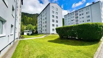 Mehrere Wohngebäude mit gepflegten Grünflächen und Hecken in bergiger Umgebung.