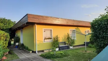 Gelbes Einfamilienhaus mit gepflegtem Vorgarten, Rasenfläche und Holzverkleidung am Dach.
