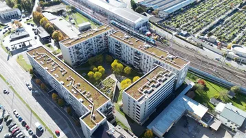 Luftaufnahme von Wohngebäuden mit begrünten Dächern, umgeben von städtischer Infrastruktur und Bergen im Hintergrund.