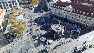 Luftaufnahme eines belebten Stadtplatzes mit Straßenbahnen, Gebäuden und vielen Menschen.