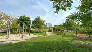 Grüner Spielplatzbereich mit Sandfläche, Holzspielgeräten und Bäumen unter hellem Himmel.