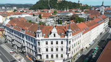 Luftaufnahme eines historischen Gebäudes an einer Straßenecke mit Blick auf die Stadt und den Schlossberg.