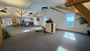 Geräumiges Großraumbüro mit hohen Decken, sichtbaren Holzbalken und modernen Ringlampen, ausgestattet mit Teppichboden und viel Tageslicht.