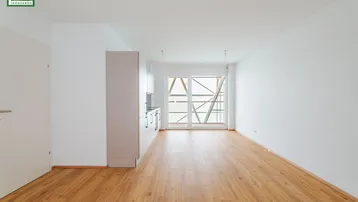 Helles, leeres Zimmer mit integrierter Küchenzeile und Zugang zu einem Balkon.