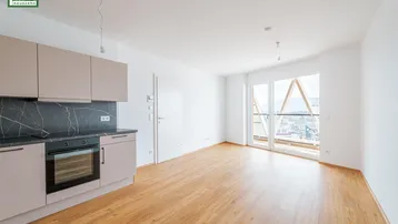 Moderne Einbauküche mit dunkler Arbeitsplatte und hellem Laminatboden, angrenzend an einen hellen Wohnbereich mit Balkonzugang.