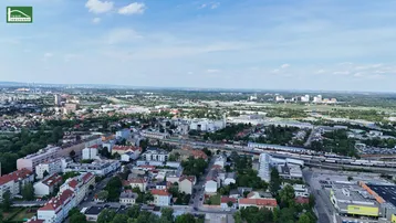 Weitläufige Luftaufnahme einer Stadtlandschaft mit Wohngebäuden, Grünflächen und Infrastruktur unter blauem Himmel.