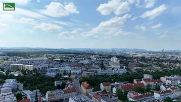 Weitläufige Luftaufnahme einer Stadt mit Wohngebäuden, Grünflächen und Bergen im Hintergrund unter blauem Himmel.