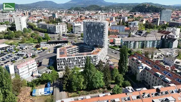 Panoramablick auf den modernen Gebäudekomplex im städtischen Umfeld mit Bergen im Hintergrund und umliegender Bebauung.