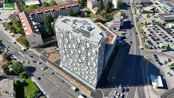 Luftaufnahme eines modernen Gebäudes mit markanter Fassade und Solaranlage auf dem Dach, umgeben von städtischer Infrastruktur.