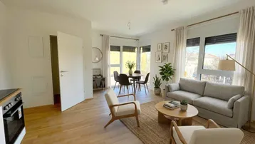 Helles, modern eingerichtetes Wohn-Esszimmer mit offener Küche, Parkettboden und großen Fenstern.