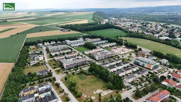 Weitwinkelaufnahme der Wohnsiedlung, umgeben von Feldern, Wäldern und Bergen am Horizont.