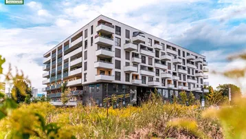 Modernes Mehrfamilienhaus mit vielen Balkonen, umgeben von grüner Vegetation unter bewölktem Himmel.