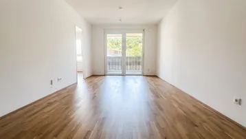 Helles, leeres Zimmer mit Holzboden und Zugang zu einem Balkon mit Blick ins Grüne.