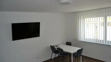 Heller Raum mit einem Tisch, vier Stühlen und einem Wandfernseher, ideal als Büro oder Besprechungszimmer.
