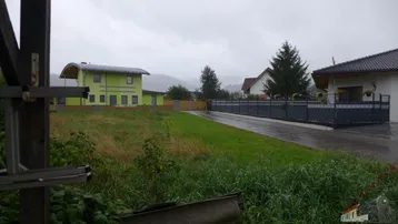 Blick auf ein modernes grünes Haus mit Solaranlage und angrenzendem Grundstück an einem bewölkten Tag.