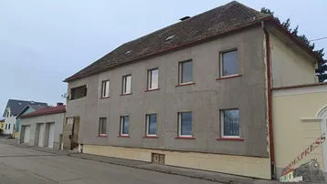 Graues Mehrfamilienhaus mit mehreren Fenstern und einem Satteldach an einer ruhigen Straße.