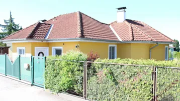 Gelbes Einfamilienhaus mit rotem Ziegeldach, umgeben von einem grünen Zaun und Hecke.