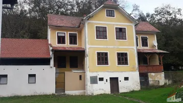 Historisches gelbes Gebäude mit rotem Ziegeldach, Holzbalkon und umgebender Natur.