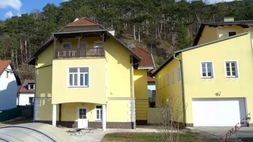 Gelbes Wohnhaus mit Balkon, Garage und Gartenbereich vor bewaldetem Hügel.