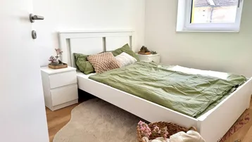 Helles Schlafzimmer mit weißem Bett, grüner Bettwäsche und Holzfußboden, ideal für erholsame Nächte.