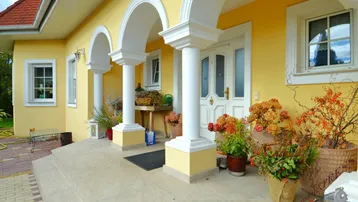 Eingangsbereich des Hauses mit gelber Fassade, weißen Säulen und dekorativen Pflanzen.