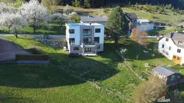 Luftaufnahme eines modernen Hauses mit Flachdach und Solaranlage, eingebettet in eine grüne Hügellandschaft.