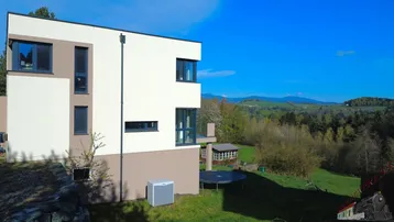 Modernes Einfamilienhaus mit weißer und beiger Fassade, umgeben von grüner Landschaft und Hügeln.