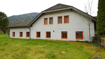 Weißes Haus mit orangefarbenen Fensterrahmen auf einem grünen Hügel mit Bergblick.