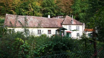 Ländliches Haus mit traditionellem Ziegeldach, umgeben von üppiger Vegetation und Bäumen.