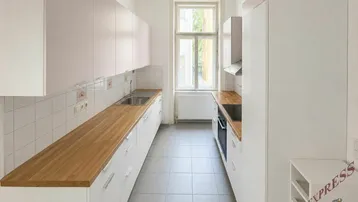 Helle Küche mit weißen Schränken, Holz-Arbeitsplatten und Fliesenboden, gut beleuchtet durch ein Fenster.