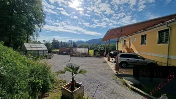 Weitläufiger Kiesparkplatz vor dem gelben Gebäude mit einem Gewächshaus und Bergblick im Hintergrund.