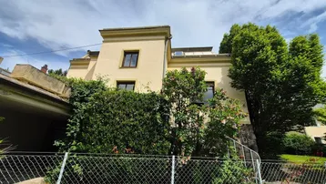 Gelbe Fassade eines Hauses mit üppiger Hecke und Einfahrt unter blauem Himmel.