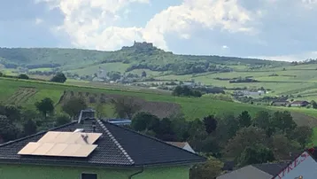 Blick auf die grüne Landschaft mit Weinbergen und einer Burg in der Ferne, im Vordergrund ein Haus mit Solaranlage.