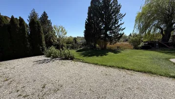 Großer Gartenbereich mit Rasenfläche, hohen Bäumen und einem Kiesparkplatz unter blauem Himmel.