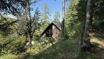 Das rustikale Holzhaus liegt idyllisch eingebettet in eine bewaldete Hügellandschaft unter blauem Himmel.