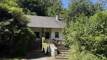 Traditionelles Landhaus umgeben von üppigem Grün und hohen Bäumen, mit einer Steintreppe zum Eingang.