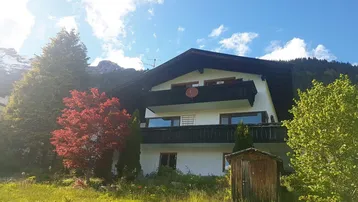 Traditionelles Haus in den Bergen mit Balkonen und umgebender Natur unter blauem Himmel.