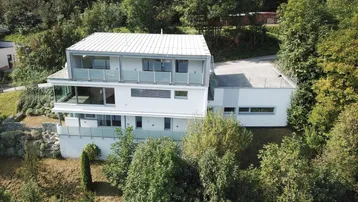 Moderne Architektur eines Mehrfamilienhauses in Hanglage, umgeben von üppiger grüner Vegetation.