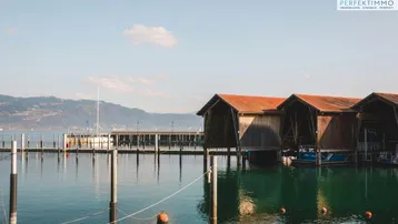 Bootehäuser am See mit Bergen im Hintergrund unter klarem Himmel, umgeben von ruhigem Wasser.
