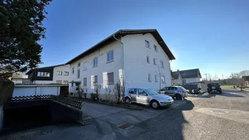 Mehrfamilienhaus mit weißer Fassade, Parkplätzen und Zufahrt zu einer Tiefgarage unter blauem Himmel.