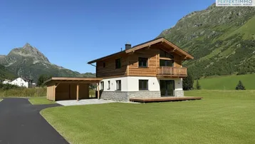 Modernes Holzhaus mit Carport, Terrasse und Bergblick in grüner alpiner Landschaft.
