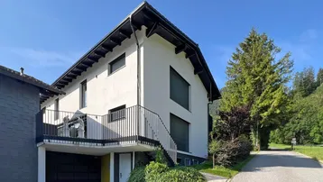 Modernes Wohnhaus mit Balkon und Garage, umgeben von grüner Natur und Bergen.