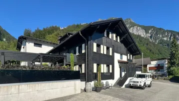 Traditionelles Holzhaus mit dunkler Fassade und Bergblick unter blauem Himmel.