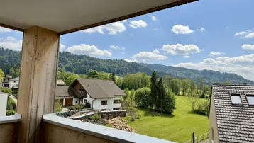Balkon mit Holzboden und weitem Blick auf grüne Hügel und bewaldete Berge unter blauem Himmel.