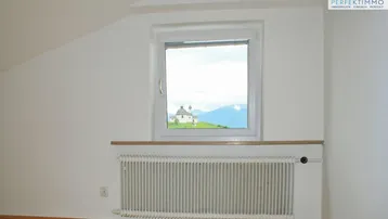 Dachzimmer mit Holzboden, weiß gestrichenen Wänden und Fenster mit Fernblick auf eine Kirche.