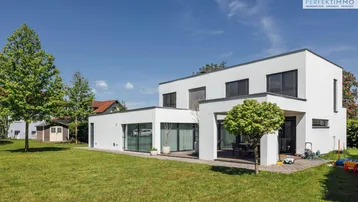 Modernes Einfamilienhaus mit weißer Fassade, großem Rasen und kleiner Gartenhütte unter blauem Himmel.