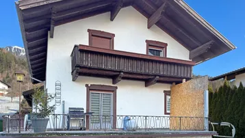 Traditionelles Haus mit Balkon und einer Terrasse im Erdgeschoss, umgeben von alpiner Landschaft.